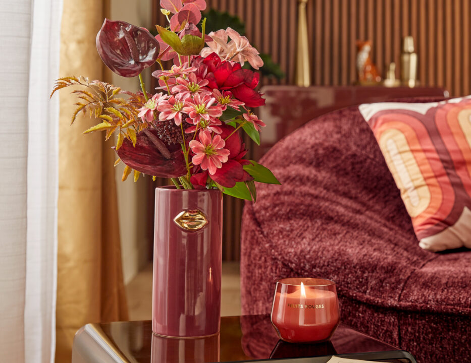 Vase bordeaux rempli de fleurs sur table basse près d'un fauteuil dans les mêmes tons.