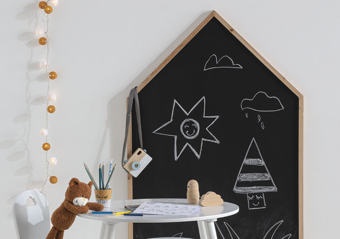 pizarrón infantil en forma de casa con dibujos de tiza al lado de una mesa de actividades y suministros creativos